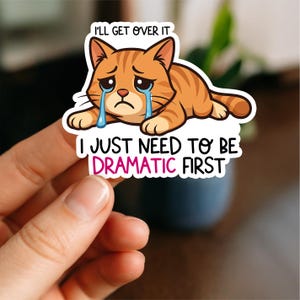 May include: A sticker showing a cartoon orange tabby cat with tears, and the text "I'll get over it" and "I just need to be dramatic first". The cat is lying down. The sticker is white with black and pink text.