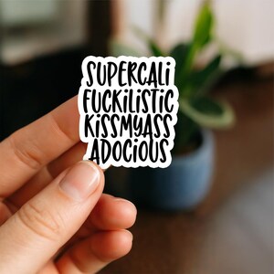 Könnte beinhalten: Ein weißer Aufkleber mit schwarzem Text, der "SUPERCALI FUCKILISTIC KISSMYASS ADOCIOUS" lautet. Der Aufkleber wird von einer Hand gehalten, mit einem verschwommenen Hintergrund einer Pflanze in einem Topf.