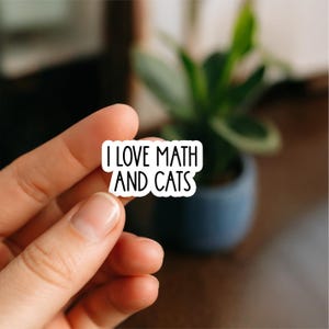 May include: A white sticker with black text that reads "I LOVE MATH AND CATS." The sticker is held between fingers. The background is blurred, with a green plant in a blue pot visible.