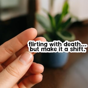 May include: A white sticker with black text that reads "flirting with death... but make it a shift." The sticker is held in a person's hand, with a blurred background of a plant in a dark blue pot.