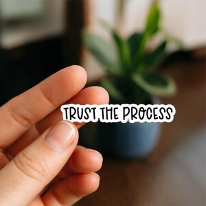 May include: A white sticker with the phrase "TRUST THE PROCESS" in black, handwritten-style lettering. The sticker is held between fingers, with a blurred background featuring a plant in a dark blue pot.