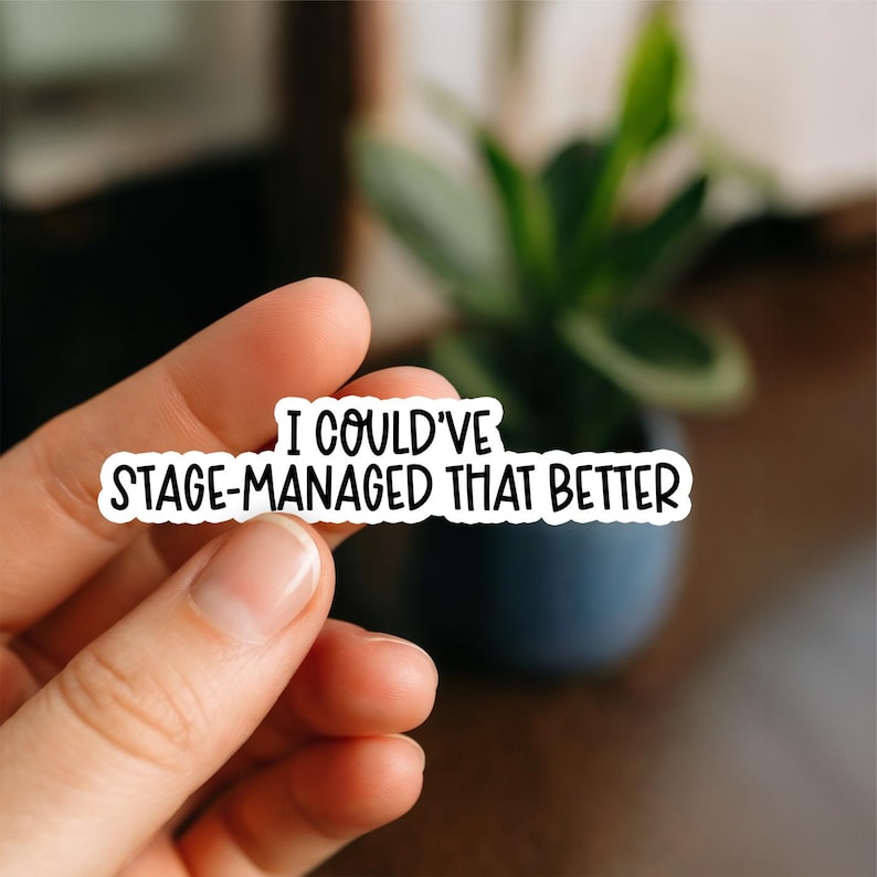 May include: A white sticker with black text that reads "I COULD'VE STAGE-MANAGED THAT BETTER". The sticker is held between fingers, with a blurred background of a plant in a dark blue pot.