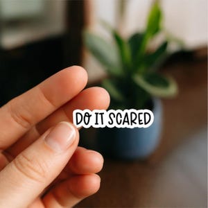 May include: A white sticker with the text "DO IT SCARED" in black. The sticker is held in front of a blurred background with a potted plant. The sticker has a black border.