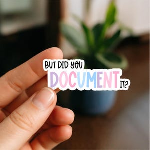 May include: A colorful sticker with the text "BUT DID YOU DOCUMENT IT?" in a playful font. The word "DOCUMENT" is in a gradient of pink, blue, and purple. The sticker is held by a hand, with a blurred background of a plant and dark surface.