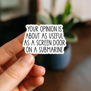 May include: A white sticker with black text: "YOUR OPINION IS ABOUT AS USEFUL AS A SCREEN DOOR ON A SUBMARINE." The sticker is held by a hand, with a blurred background.