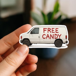 May include: A white van sticker with the text "FREE CANDY" in red, dripping lettering. The van has black wheels and a side window. The sticker is held by a hand.
