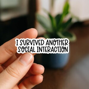 Könnte beinhalten: Ein weißer Aufkleber mit dem schwarzen Text "I SURVIVED ANOTHER SOCIAL INTERACTION". Der Aufkleber wird von einer Hand gehalten, mit einem verschwommenen Hintergrund mit einer Pflanze in einem blauen Topf.