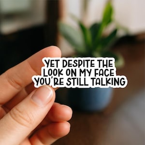 Könnte beinhalten: Ein weißer Aufkleber mit schwarzem Text, der lautet: "YET DESPITE THE LOOK ON MY FACE YOU'RE STILL TALKING". Der Aufkleber wird von einer Hand gehalten, mit einem verschwommenen Hintergrund einer Pflanze und einem dunkelblauen Topf.