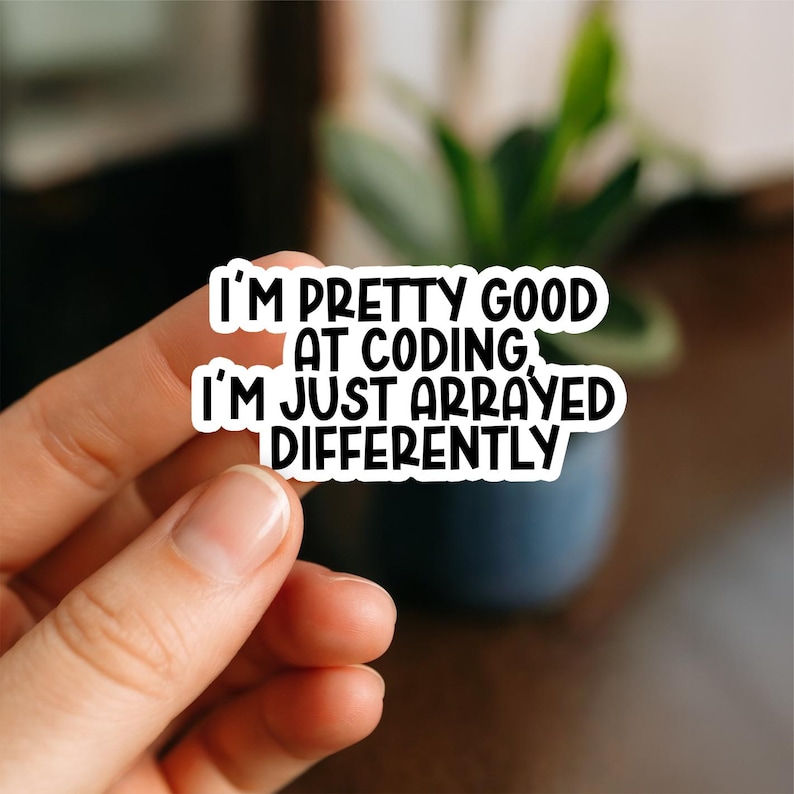May include: A white sticker with black text that reads "I'M PRETTY GOOD AT CODING, I'M JUST ARRAYED DIFFERENTLY." The sticker is held by a hand, with a blurred background of a plant and a dark surface.