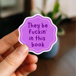 May include: A purple sticker with a white border and the text "They be fuckin' in this book". The sticker is held by a hand, with a blurred background of a plant.