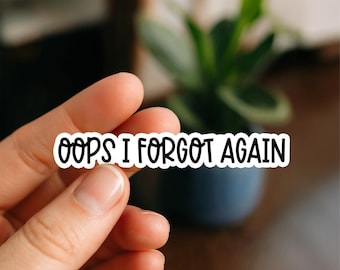May include: A white sticker with black text that reads "OOPS I FORGOT AGAIN". The sticker is held between two fingers. A blurred background shows a plant in a dark blue pot.