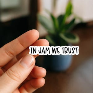 May include: A white sticker with the text "IN JAM WE TRUST" in black, held by a hand. The sticker is rectangular and has a black border. The background is blurred, with a plant in a dark blue pot.
