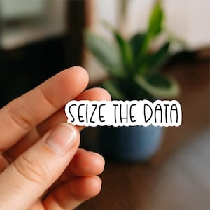 May include: A white sticker with the words "SEIZE THE DATA" in black, sans-serif font. The sticker is held by a hand, with a blurred background featuring a plant in a dark blue pot.