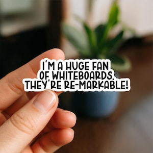 May include: A white sticker with black text that reads "I'M A HUGE FAN OF WHITEBOARDS. THEY'RE RE-MARKABLE!" The sticker is held by a hand, with a blurred background of a plant and a dark surface.