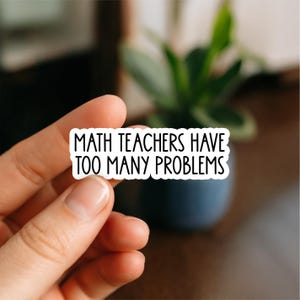 May include: A white sticker with black text that reads "MATH TEACHERS HAVE TOO MANY PROBLEMS." The sticker is held between fingers against a blurred background of a plant and a dark blue pot.