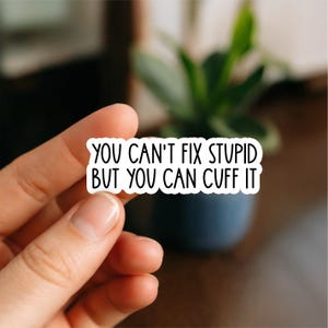 May include: A white sticker with black text that reads "YOU CAN'T FIX STUPID BUT YOU CAN CUFF IT." The sticker is held by a person's fingers, with a blurred background of a plant in a blue pot.