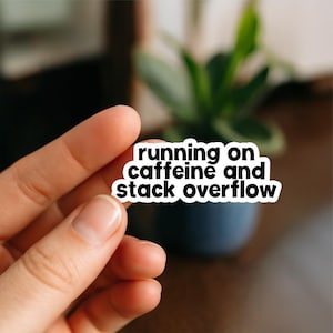 Peut inclure: Un autocollant blanc avec du texte noir qui dit "running on caffeine and stack overflow". L'autocollant est tenu par une main, avec un arrière-plan flou d'une plante dans un pot bleu foncé.