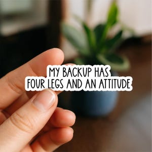 May include: A white sticker with black text that reads "MY BACKUP HAS FOUR LEGS AND AN ATTITUDE." The sticker is held by a person's hand, with a blurred background of a plant and a dark blue pot.