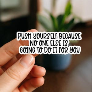 May include: A white sticker with black text that reads "PUSH YOURSELF, BECAUSE NO ONE ELSE IS GOING TO DO IT FOR YOU." The sticker is held in a person's hand, with a blurred background of a plant and a dark blue pot.