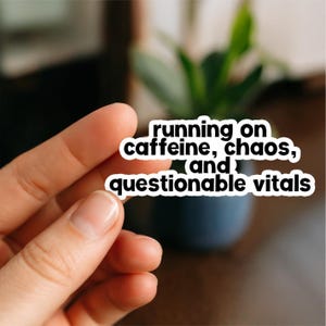 May include: A white sticker with black text that reads "running on caffeine, chaos, and questionable vitals." The sticker is held between two fingers. A blurred background includes a plant and a dark blue pot.