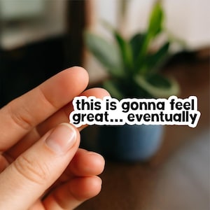 May include: A white sticker with black text that reads "this is gonna feel great... eventually". The sticker is held in a person's hand, with a blurred background of a plant in a dark blue pot.
