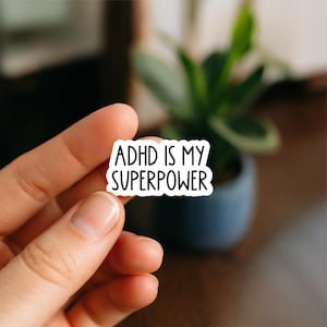 May include: A white sticker with the text "ADHD IS MY SUPERPOWER" in a bold, black font. The sticker is held between fingers, with a blurred background featuring a green plant in a dark blue pot.