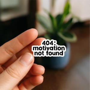 Peut inclure: Un autocollant blanc avec le texte noir "404: motivation not found". L'autocollant est tenu par une main, avec un arrière-plan flou d'une plante dans un pot bleu foncé.