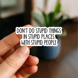 May include: A white sticker with black text that reads "DON'T DO STUPID THINGS IN STUPID PLACES WITH STUPID PEOPLE." The sticker is held by a person's fingers, with a blurred background of a plant and a dark blue pot.