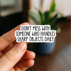 Può includere: Un adesivo bianco con testo nero che recita "DON'T MESS WITH SOMEONE WHO HANDLES SHARP OBJECTS DAILY." L'adesivo è tenuto in mano, con uno sfondo sfocato di una pianta e un vaso blu scuro.