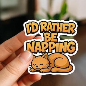 May include: A sticker with the text "I'd Rather Be Napping" in orange and black. Below the text is an orange cartoon cat curled up and sleeping. The sticker has a white border.