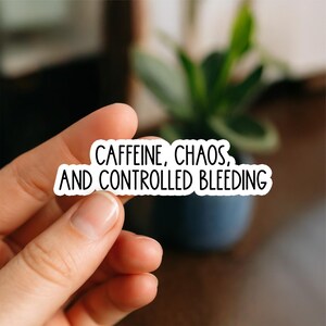 May include: A white sticker with black text that reads "CAFFEINE, CHAOS, AND CONTROLLED BLEEDING." The sticker is held between two fingers. A blurred background includes a small potted plant.