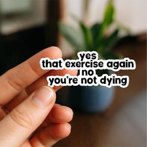 May include: A white sticker with black text that reads "yes that exercise again no you're not dying." The sticker is held between fingers. A blurred background includes a plant and a dark blue pot.
