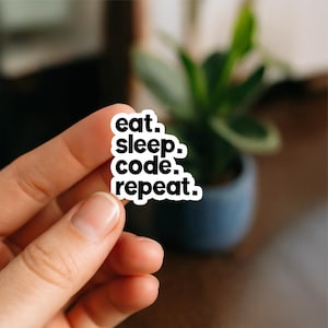 Può includere: Un adesivo bianco con testo nero che recita "eat. sleep. code. repeat." L'adesivo è tenuto tra le dita, con uno sfondo sfocato di una pianta in vaso.