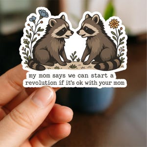 May include: A sticker showing two cartoon raccoons. The raccoons are brown and grey with black masks. The sticker has the text: "my mom says we can start a revolution if it's ok with your mom."