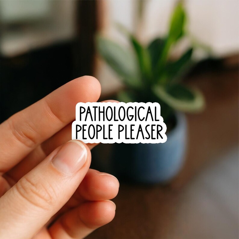 Puede incluir: Una pegatina blanca con el texto "PATHOLOGICAL PEOPLE PLEASER" en negro, sostenida entre los dedos. La pegatina tiene un borde negro. El fondo est&aacute; borroso, con una planta en una maceta azul oscuro.
