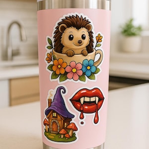 A pink tumbler with various stickers on it, including a hedgehog, a fairy house, and a vampire-like character.