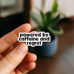 Può includere: Un adesivo bianco con testo nero che recita "powered by caffeine and regret". L'adesivo è tenuto in mano, con uno sfondo sfocato di una pianta in un vaso blu scuro.