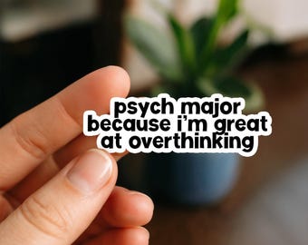 May include: A white sticker with black text that reads "psych major because I'm great at overthinking." The sticker is held in a person's hand, with a blurred background of a plant in a blue pot.