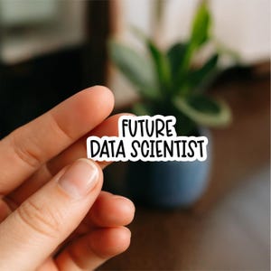 May include: A white sticker with the words "FUTURE DATA SCIENTIST" in black, bold lettering. The sticker is held between fingers, with a blurred background showing a plant in a dark blue pot.