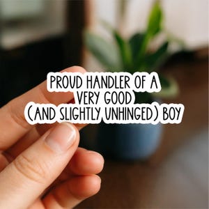 May include: A white sticker with black text that reads "PROUD HANDLER OF A VERY GOOD (AND SLIGHTLY UNHINGED) BOY." The sticker is held by a hand, with a blurred background of a plant and a dark blue pot.