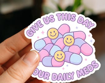 May include: A sticker featuring a cluster of cartoon pills in shades of pink, purple, and blue, each with a smiling face. The sticker has the text "GIVE US THIS DAY OUR DAILY MEDS" in a purple, rounded font.