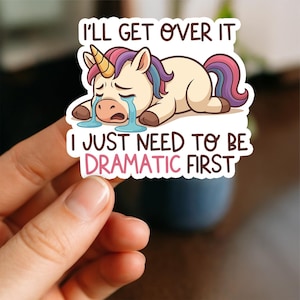 May include: A sticker featuring a cartoon unicorn with a golden horn, crying tears. The unicorn has a purple, blue, and pink mane and tail. Text on the sticker reads, "I'll get over it. I just need to be dramatic first."