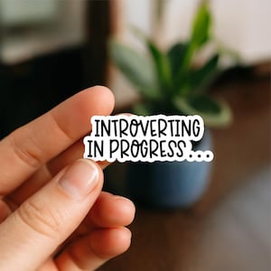 May include: A white sticker with the text "INTROVERTING IN PROGRESS..." in black, held by a hand. The sticker is rectangular with rounded corners. The background is blurred, featuring a plant and a dark blue pot.