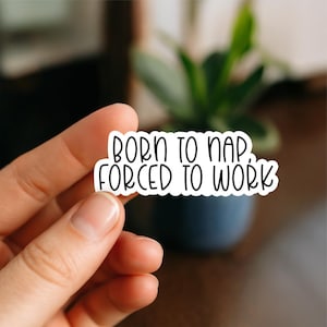 May include: A white sticker with black text that reads "BORN TO NAP, FORCED TO WORK." The sticker is held in a person's hand, with a blurred background of a plant in a dark blue pot.