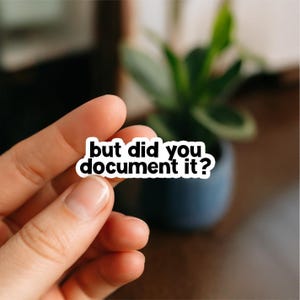 May include: A white sticker with black text that reads "but did you document it?" The sticker is held by a hand in front of a blurred background with a plant in a dark blue pot.