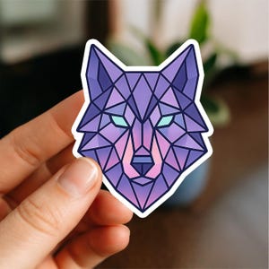 May include: A geometric sticker of a wolf's head. The design uses shades of purple, pink, and blue, with a polygonal style. The wolf's eyes are a bright teal colour. The sticker is held by a hand.