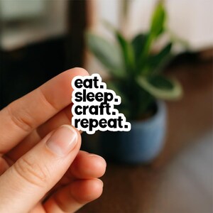 May include: A white sticker with black text that reads "eat. sleep. craft. repeat." The sticker is held between two fingers. The background is blurred, with a potted plant visible.