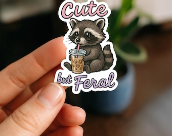 May include: A sticker featuring a cartoon raccoon drinking from a beverage cup with ice. The raccoon is brown and gray, with the text "Cute but Feral" in pink and purple script.