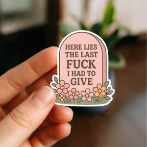 May include: A sticker with a pink tombstone design. The text on the tombstone reads "HERE LIES THE LAST FUCK I HAD TO GIVE." The tombstone is decorated with pink and orange flowers and has a white border.