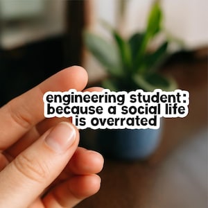 May include: A white sticker with black text: "engineering student: because a social life is overrated." The sticker is held by a hand, with a blurred background of a plant in a dark blue pot.